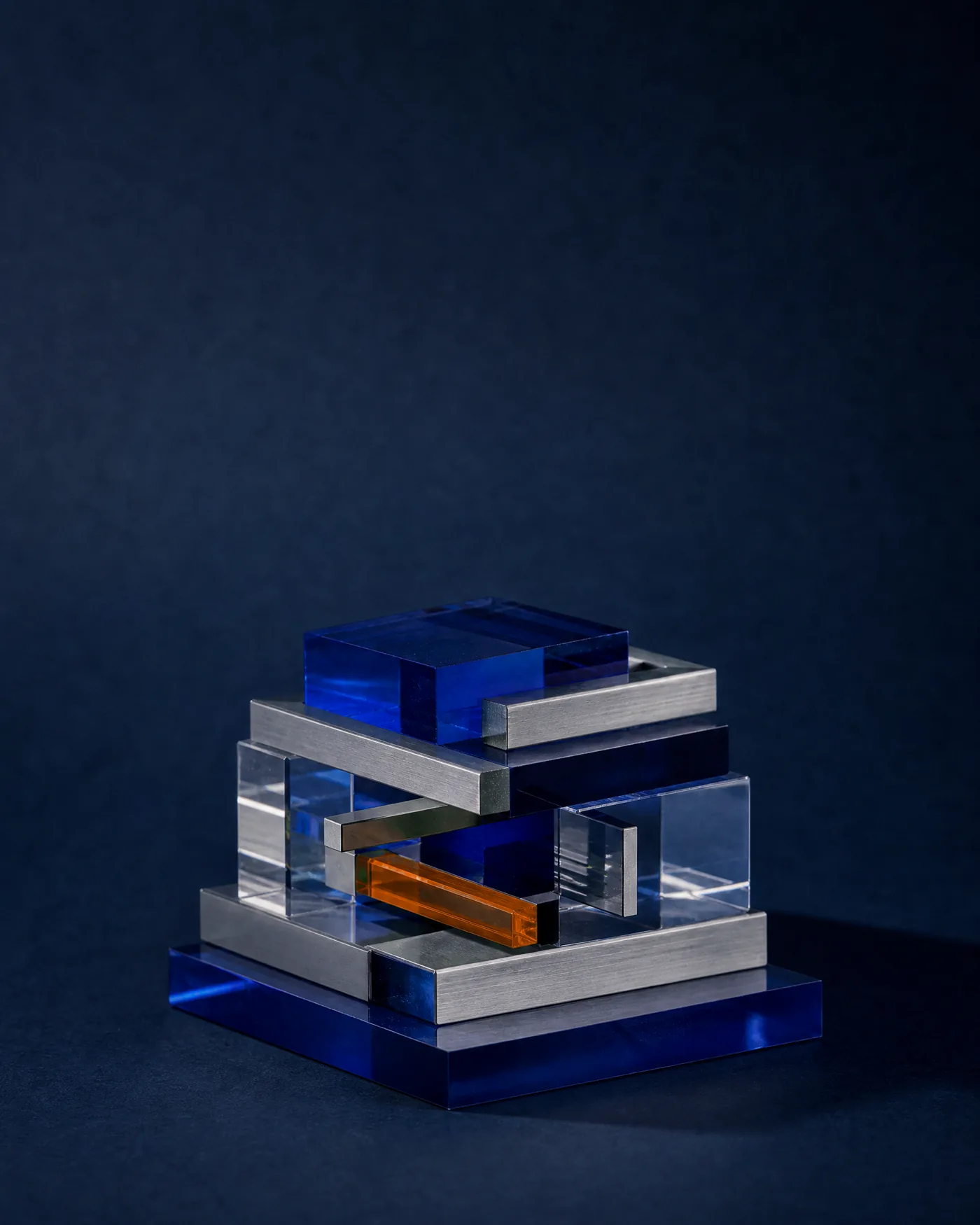 Escultura de bloques de vidrio azul y aluminio con una pieza naranja, sobre fondo azul oscuro